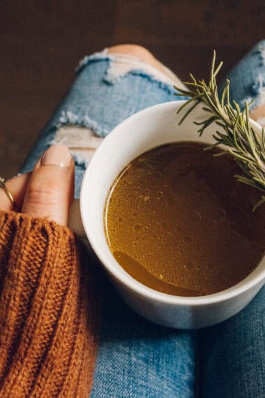a cup of bone broth with a fresh rosemary spring being held on someone's lap.