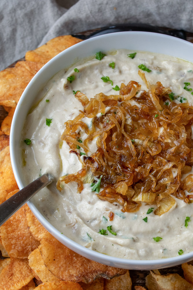 a bowl of vegan French onion dip topped with caramelized onions and chopped parsley, surrounded by potato chips.