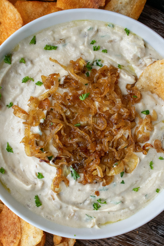 a bowl of vegan French onion dip topped with caramelized onions and chopped parsley, surrounded by potato chips.