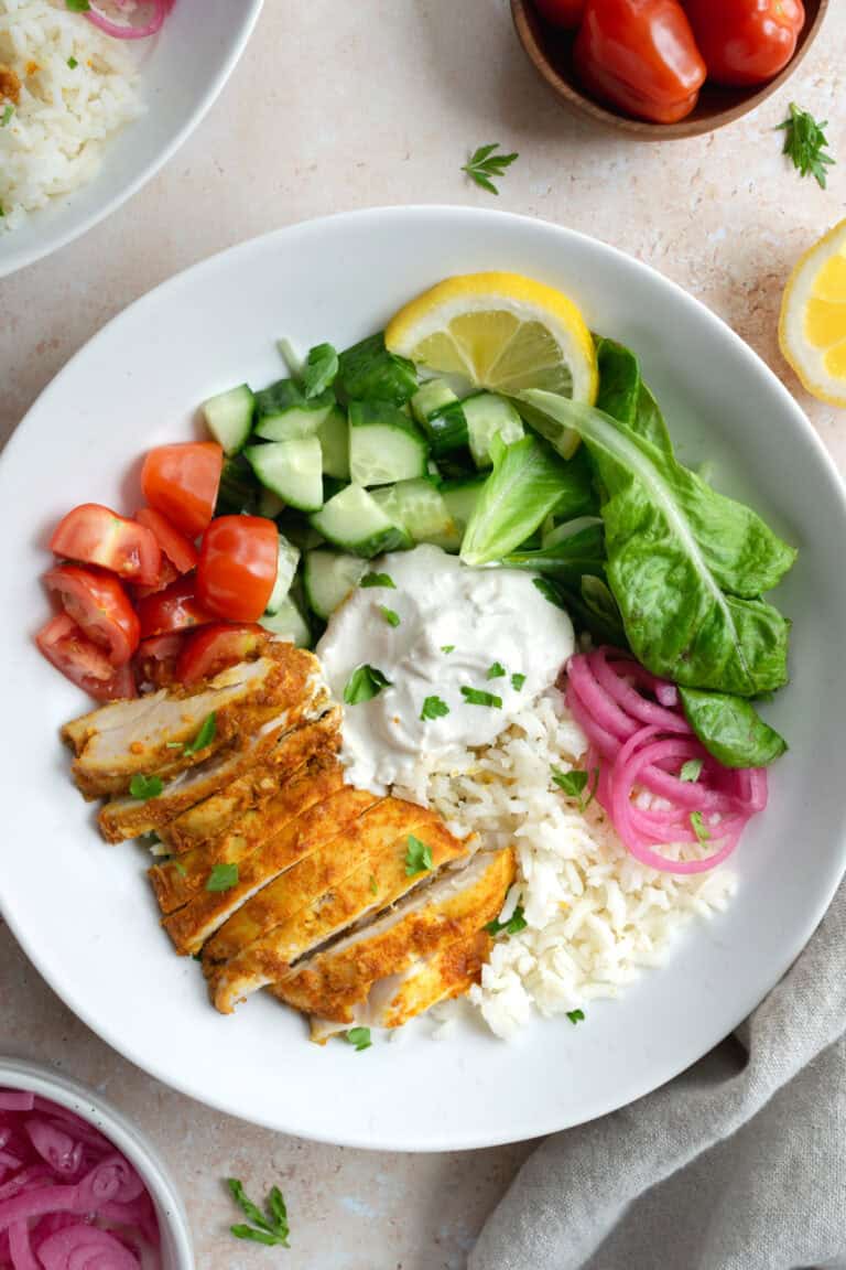 chicken shawarma bowl besides a bowl of grape tomatoes, lemon and a bowl of pickled red onions.