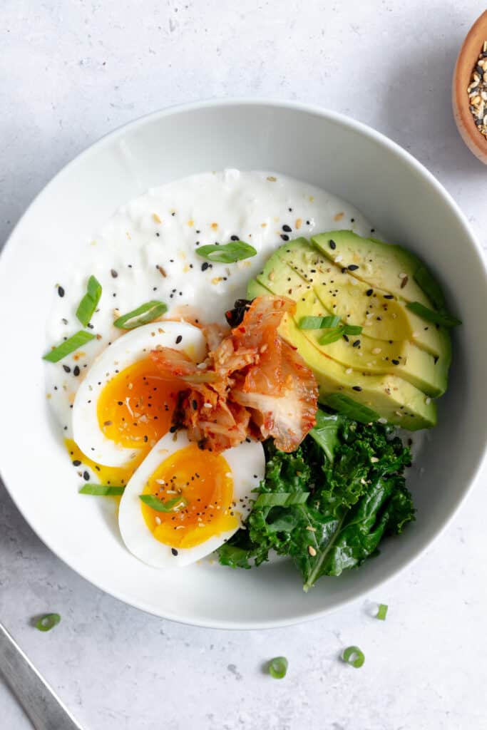 cottage cheese in a white bowl topped with egg, kale, avocado, kimchi and green onion.