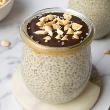 a jar of peanut butter chia pudding topped with dark chocolate, peanuts and sea salt with a bowl of peanuts and chocolate chips in the background.