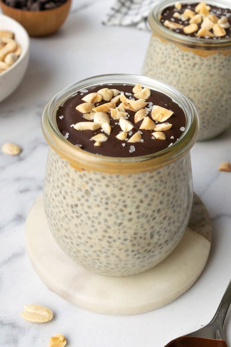 a jar of peanut butter chia pudding topped with dark chocolate, peanuts and sea salt with a bowl of peanuts and chocolate chips in the background.