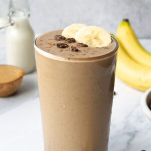 a smoothie in a tall glass topped with two slices of banana and whole coffee beans, with bananas, milk and peanut butter in the background.