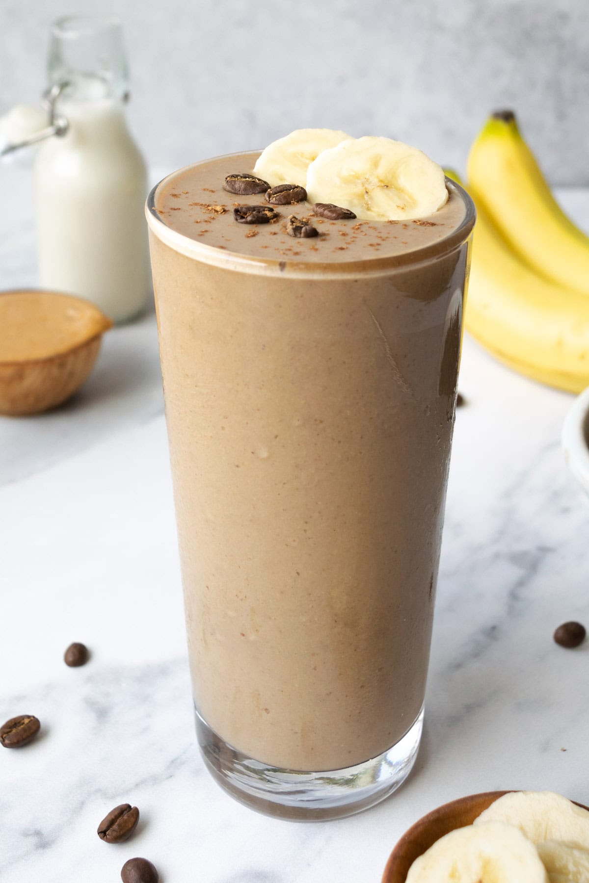 a smoothie in a tall glass topped with two slices of banana and whole coffee beans, with bananas, milk and peanut butter in the background.