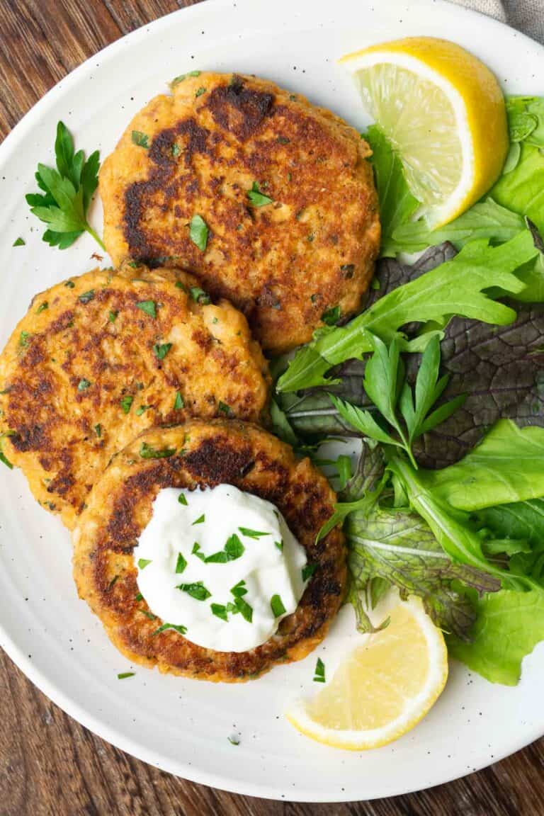 three salmon patties topped with chopped parsley and yogurt, and a green salad on a white plate.
