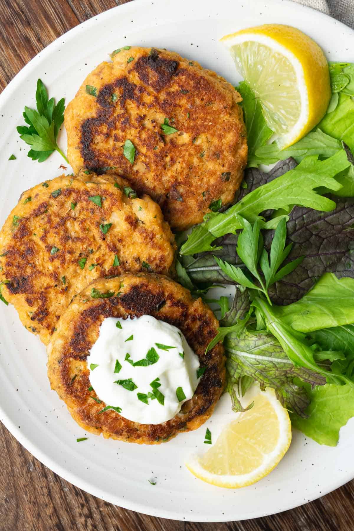 three salmon patties topped with chopped parsley and yogurt, and a green salad on a white plate.
