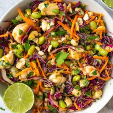 shrimp edamame salad in a white bowl beside a bowl of cashews and a bowl of cut up limes.