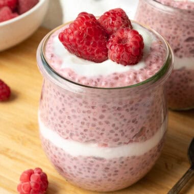 two glass jars of raspberry chia pudding topped with yogurt and fresh raspberries.