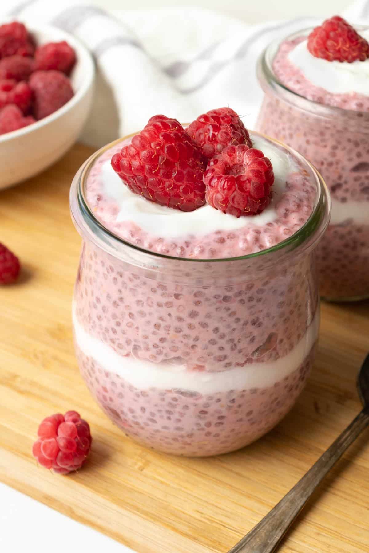 two glass jars of raspberry chia pudding topped with yogurt and fresh raspberries.
