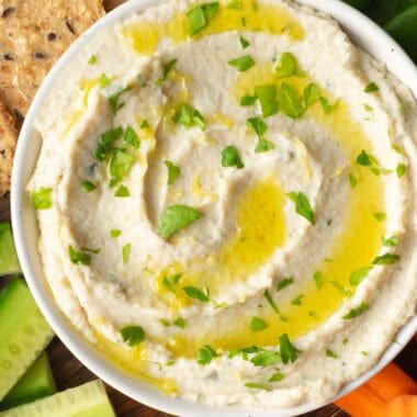 artichoke white bean dip in a white bowl surrounded by crackers, cucumber, tomatoes, carrots and peas.