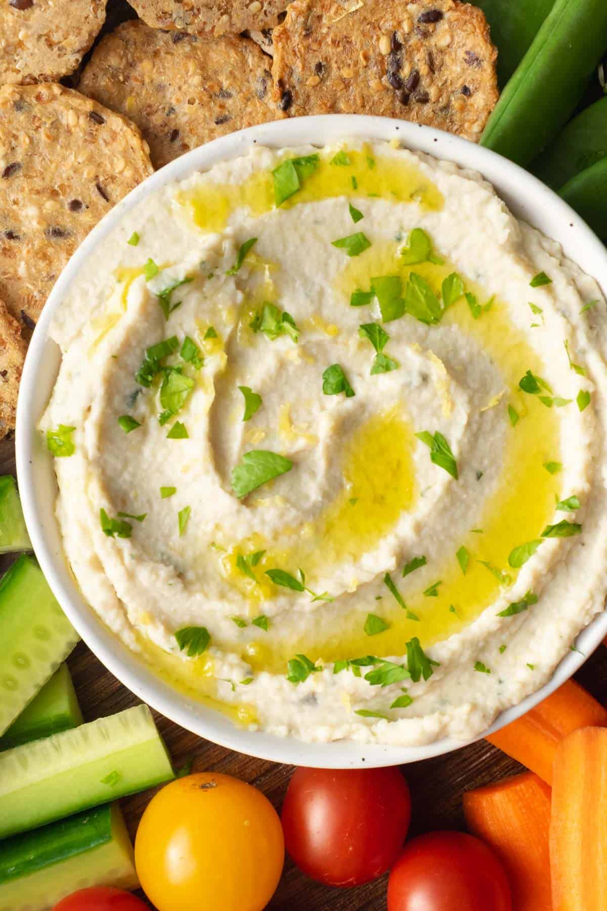 artichoke white bean dip in a white bowl surrounded by crackers, cucumber, tomatoes, carrots and peas.
